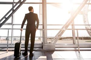 Rear view of businessman with suitcase standing next to window at airport, looking at airplane, copy space. Wealthy man in black suit waiting for departure, looking through the window, sun flare photo