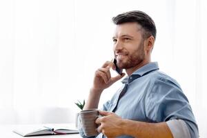 Handsome freelancer man talking on cellphone and drinking coffee at home, millennial guy sitting at desk with laptop and notepad near window, enjoying mobile communication and remote job, copy space photo