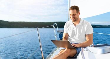 Happy Man On Yacht Sitting With Laptop Working Living On Sailboat During Summer. Liveaboard Tour. Empty Space For Text photo