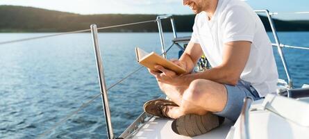 Dream Vacation. Man Reading Book Sitting On Yacht Deck Sailing Across The Sea Outdoor. Male Enjoying Summer Day On Sailboat. Yachting, Tourism Concept. Blank Space For Text, Side View photo