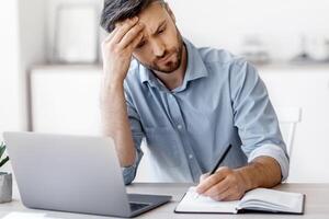 Serious Millennial Businessman Taking Notes And Working On Laptop Computer In Modern Office, Busy Entrepreneur Planning Business Schedule, Writing IN Notepad, Sitting At Desk On Workplace, Free Space photo