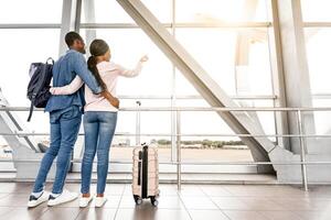 Post-Quarantine Travelling. Black Loving Couple Looking At Airport Window And Hugging, Excited About Future Flight, Rear View With Copy Space photo