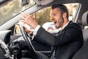 Stressed businessman driving luxury car, stuck in traffic, late to airport, side view, copy space. Furious middle-aged man in expensive suit screaming and gesturing while driving auto photo