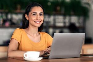 Cheerful female student using laptop at cafe, drinking coffee while studying, making report for university, copy space. Young lady sitting in front of laptop at cafeteria, smiling at camera photo