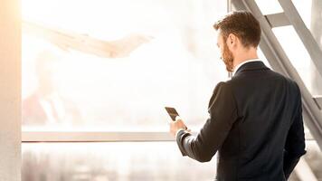 Back view of middle-aged entrepreneur waiting for departure, checking information about flight on mobile phone, bearded businessman standing in airport with luggage, panorama with copy space photo