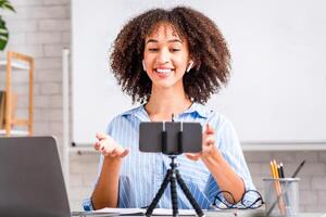 Online English lesson for students in self-isolation at home. Happy african american woman in wireless headphones looks in web camera of smartphone, sitting at table with laptop during covid-19 photo