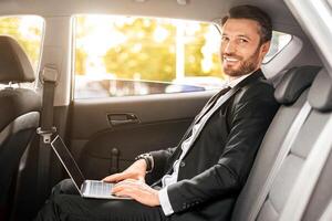 Cheerful businessman sitting in car, using laptop, working online. Handsome man in suit working on notebook while going to airport, side view. Modern technologies in business concept photo