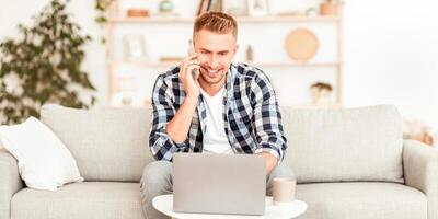 Smiling happy man using laptop and talking on mobile phone at home, having pleasant conversation with friend or client, typing, satisfied student holding smartphone, sitting on sofa in living room photo