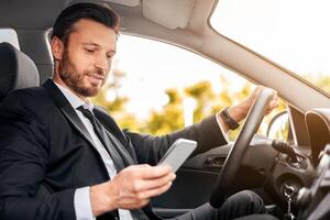 Handsome businessman driving car, using mobile phone, checking location of business meeting, side view, copy space. Bearded entrepreneur man in suit using gps navigation on smartphone photo