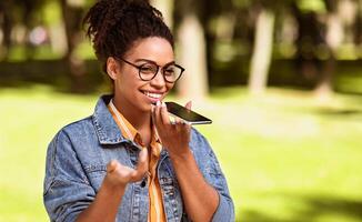 Happy Black Lady Talking On Cellphone Using Loudspeaker Mode Walking Outdoors In City Park. Smartphone User Chatting On Phone, Modern Mobile Communication Concept. Empty Space For Text photo