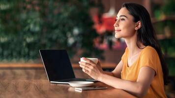 Happy young lady using laptop at cafe, drinking coffee while studying, making report for university, copy space. Young woman sitting in front of laptop at cafeteria, drinking tea and dreaming photo