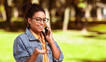 contento africano americano mujer hablando en Teléfono móvil, haciendo teléfono llamada caminando al aire libre en ciudad parque. móvil conversación, moderno comunicación concepto. blanco espacio para texto foto