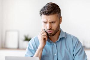 Worried Male Entrepreneur Having Problems With Laptop Computer At Work, Talking With Customer Support On Cellphone, Concerned Man Suffering Bad Internet Connection At Workplace In Office, Free Space photo