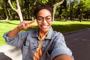 Cheerful Black Female Student Making Selfie Gesturing V-Sign Having Fun, Smiling To Camera Posing Outside Standing In Park. Self-Portrait, Mobile Photo Concept. Selective Focus