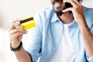 Mobile Payment. Positive Indian Man Holding Credit Card And Talking On Cellphone In Office, Transfering Money, Using Online Banking, Talking With Customer Support, Paying In Internet, Selective Focus photo