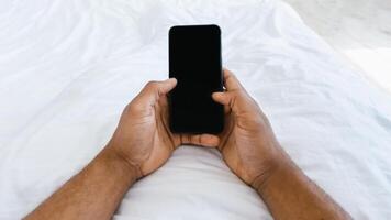 Unrecognizable African Man Using Smartphone With Blank Screen, Holding Cellphone Lying In Bed At Home. Mobile Application, Texting On Phone, And Online Chat Concept. Panorama, Cropped, POV photo