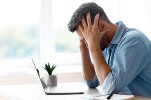 Stressed male freelancer sitting at desk in home office, touching head, having business problems or lack of inspiration while working on online project with laptop, side view with copy space photo