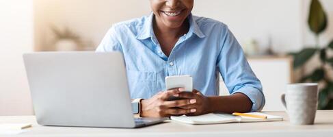 Smiling young african american businesswoman using smartphone at workplace in office, texting, browsing internet or checking new app, copy space photo