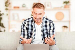 Online Communication Concept. Portrait of young man making virtual call on laptop, talking, looking at webcam and gesturing. Man chatting using computer, sitting on the couch at home photo