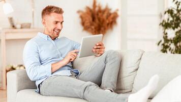 Technology Concept. Portrait of smiling man using digital tablet sitting on the couch at his modern apartment, copy space. Happy guy reading news, browsing internet, resting on sofa in living room photo