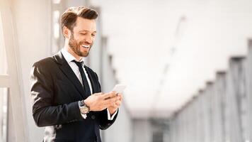 Cheerful businessman in stylish suit chatting with lover while waiting for flight in airport, using mobile phone, copy space. Happy entrepreneur got good news, reading emails from business partners photo