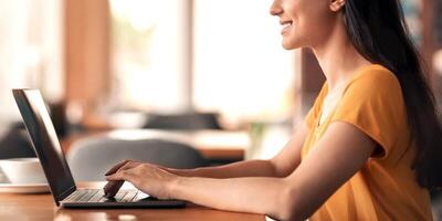 Side view of young smiling arab woman using laptop while sitting at cafe, empty space. Female freelancer working online, typing on keyboard, sending email to client, drinking coffee photo