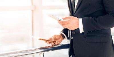 Closeup of unrecognizable businessman using phone, holding passport and flight tickets, standing by window at airport, waiting for departure, copy space. Man in suit checking email on mobile phone photo