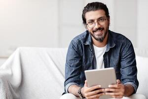 Handsome Indian Guy Holding Digital Tablet Computer And Smiling At Camera, Sitting On Couch At Home, Using Modern Technologies For Leisure, Browsing Internet Or Checking Social Networks, Free Space photo