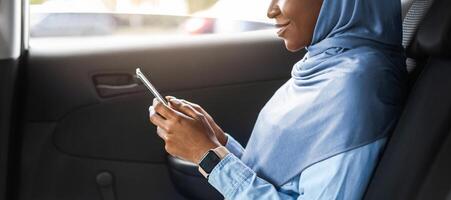 Smiling Black Muslim Female Entrepreneur Browsing Smartphone While Riding Car On Backseat, Using Taxi Services, Going To Office, Messaging On Cellphone, Answering Emails, Side View With Free Space photo