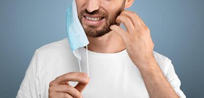 Bearded man moved protective medical mask from face to scratch beard, grey studio background, free space. Do not touch your face, avoid touching your face during pandemic, stay safe during lockdown photo
