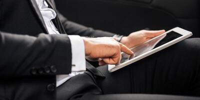 Modern technologies in business. Cropped of businessman in suit sitting in car, using digital tablet, working online. Unrecognizable man working on digital pad while going to airport, side view photo