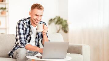 Remote Work And Studying Concept. Portrait of smiling guy typing on laptop keyboard at home in living room, sitting on the sofa, drinking coffee and using personal computer at home, copy space photo