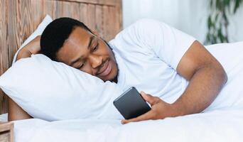 African american guy with cellphone lying in bed using mobile phone application at home. Guy looking at smartphone waiting for call resting in bedroom photo