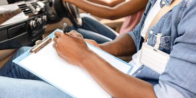 Unrecognizable black man driving instructor taking notes at chart during exam, closeup. Cropped of african american woman attending driving school, passing test, cropped, side view photo