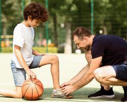 Side view of smiling adult dad helping his son to tie shoelaces, kid playing on basketball field photo