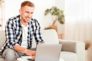 Home Office Concept. Portrait of smiling man working online on laptop sitting on the sofa and wearing wireless earbuds, copy space. Guy listening to music or podcast using computer photo