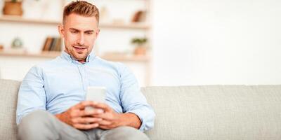 Free Time Concept. Portrait of handsome man using his smartphone, sitting on the couch at home, blurred background, copy space. Guy chatting online, browsing social media, texting internet message photo