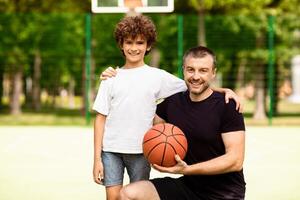 Active Lifestyle Concept. Happy boy posing with his coach on basketball field, man holding ball and looking at camera photo