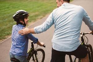 Healthy Family Relationship. Rear back view portrait of smiling father and son riding bicycles down the street in public park, man tapping and patting boy on the shoulder, teaching and showing support photo