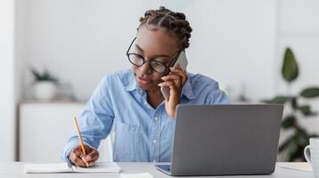 Busy Black Female Office Worker Talking On Cellphone With Client And Taking Notes, Sitting At Workplace In Modern Office, Working On Desk With Laptop, Writing Down Information To Notepad, Copy Space photo