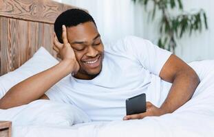 Cheerful African Man With Smartphone Browsing Internet Or Using Mobile Phone App Lying In Bed At Home. Cellphone App Advertisement, Weekend Morning Leisure Concept photo