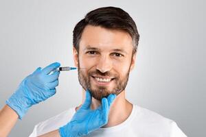 Cosmetician hands in protective gloves injecting face filler for middle-aged man over grey studio background, copy space. Smiling man getting anti-aging procedure in beauty salon or clinic photo