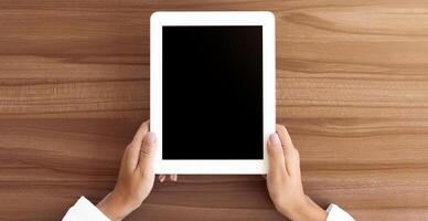 Mock up image of two hands holding digital tablet with isolated black screen, wooden table in the background photo