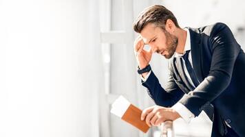 Stressful businessman waiting at airport after flight delays and cancellations, leaning on fence, holding passport with flight tickets, empty space. Entrepreneur is late for business meeting photo
