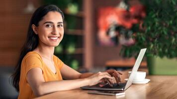 Smiling pretty young arab woman using laptop at cafe, working online, empty space. Side view of cheerful lady freelancer typing on laptop, sending email or chatting with clients, drinking tea photo
