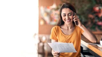 Happy arab lady having conversation on mobile phone at cafe, holding documents, working online, copy space. Smiling woman talking on smartphone with clients while drinking coffee, looking at papers photo