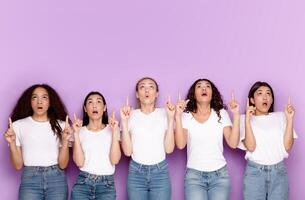 Five Surprised Diverse Ladies Pointing Fingers Upward Showing Free Space For Text Above Their Heads Posing Over Purple Background. Studio Shot. Look Up At This Offer Concept photo