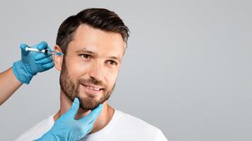 Handsome bearded man getting under eye injections in beauty clinic, looking at copy space, grey studio background. Middle-aged smiling man attending beautician, having face filler photo