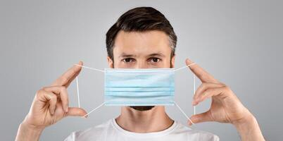 Bearded man holding medical protective mask on grey studio background. Man in white t-shirt with blue protective medical mask, healthcare concept, stay safe and healthy during pandemic photo