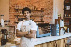 Reopening of small businesses and returning to normal after covid-19 quarantine. Smiling millennial african american guy in apron near bar with crossed arms, looking at camera, waiting for customers photo